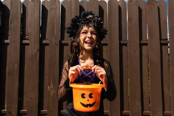 Excitée fille en costume de sorcière et couronne noire tenant seau halloween à l'extérieur — Photo de stock