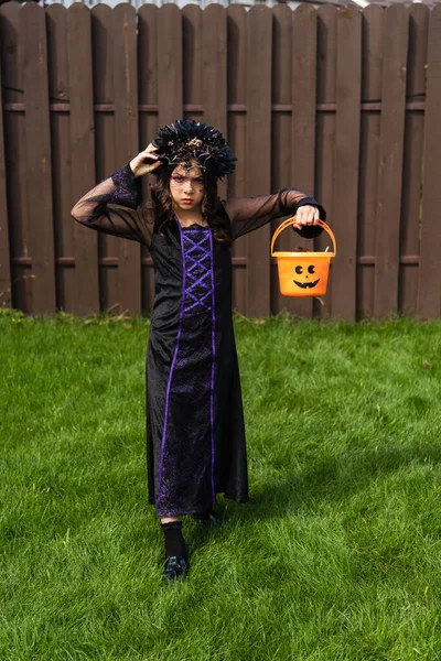 Chica seria en vestido negro y corona sosteniendo cubo de Halloween mientras camina en el patio trasero - foto de stock