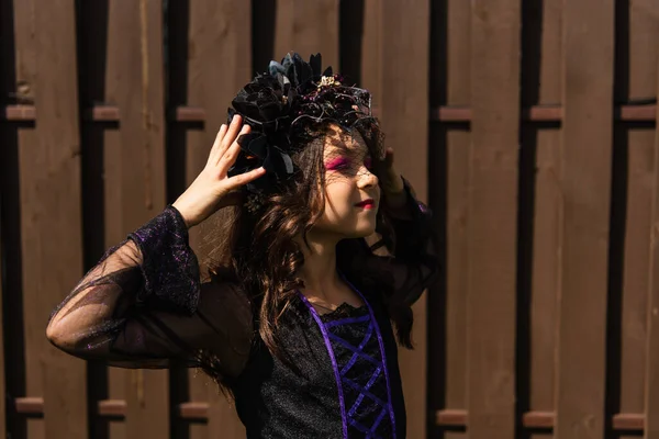 Complacido chica en bruja halloween traje tocando negro corona al aire libre - foto de stock