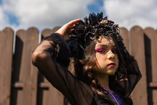 Girl in witch costume and black wreath with veil looking at camera outdoors — Stock Photo