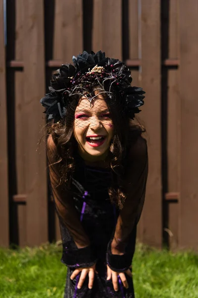 Fröhliches Mädchen in Halloween-Kostüm und schwarzem Kranz, das draußen vor der Kamera lacht — Stockfoto