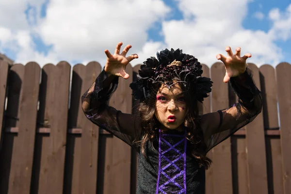Girl in witch halloween costume and black wreath showing frightening gesture — Stock Photo