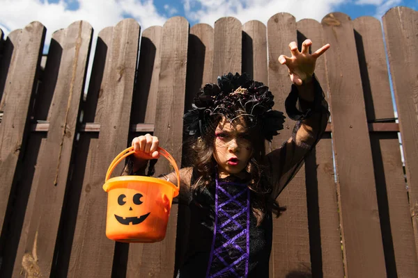 Girl in witch costume showing frightening gesture while holding trick or treat bucket — Stock Photo
