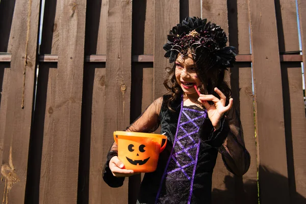 Chica en traje de bruja mirando hacia otro lado y comer regaliz palo mientras sostiene halloween cubo - foto de stock