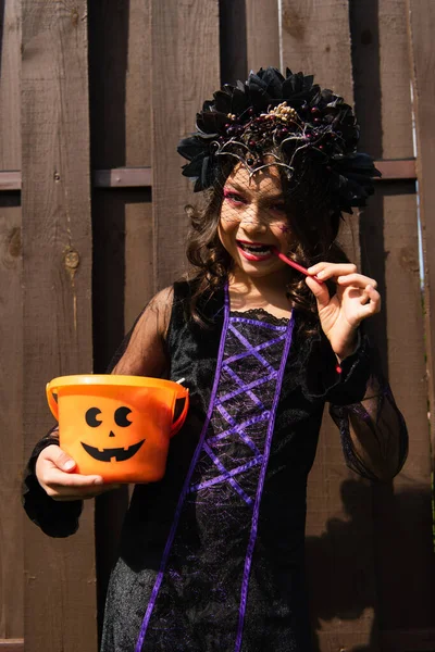 Chica en traje de bruja celebración de halloween cubo y comer regaliz palo - foto de stock