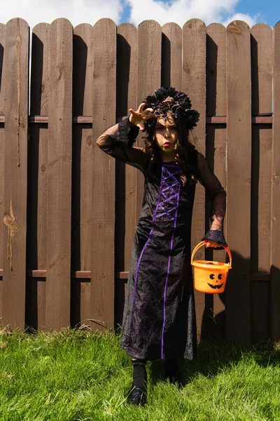 Chica en traje de bruja celebración de halloween cubo y mostrando gesto de miedo - foto de stock