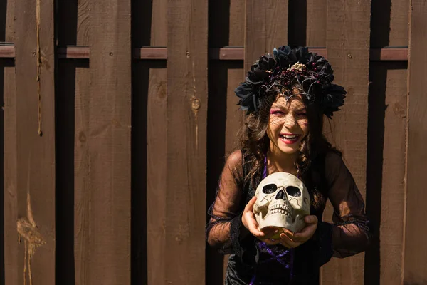 Cheerful girl in black wreath holding spooky skull and smiling at camera near wooden fence — Stock Photo