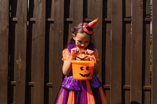 Smiling girl in party cap and clown costume looking into halloween bucket near wooden fence — Stock Photo