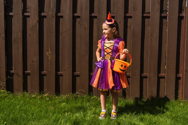 Piena lunghezza di ragazza in costume da clown in piedi con secchio di Halloween e guardando altrove — Foto stock