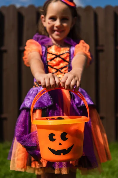 Selective focus of trick or treat bucket in hands of girl in clown costume smiling on blurred background — Stock Photo