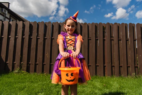 Chica en traje de payaso y gorra de fiesta de pie con cubo de Halloween en el patio trasero - foto de stock