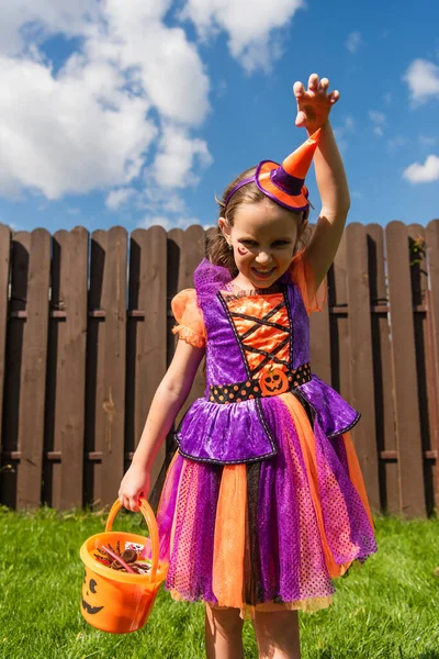 Girl in clown costume showing scary grimace and frightening gesture while holding halloween bucket — Stock Photo