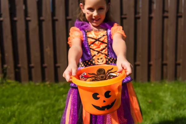 Chica borrosa en traje de payaso celebración de cubo de Halloween con araña de juguete y dulces - foto de stock