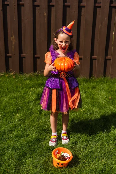 Aufgeregtes Mädchen im Clownskostüm mit Kürbis in der Nähe von Halloween-Eimer mit Süßigkeiten — Stockfoto