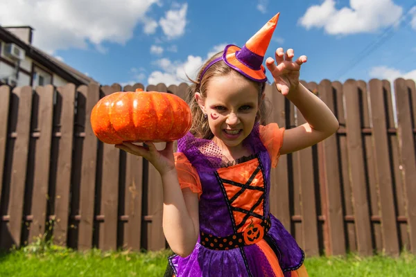 Chica en traje de payaso celebración de calabaza mientras que hace mueca de miedo y el gesto en el patio trasero - foto de stock