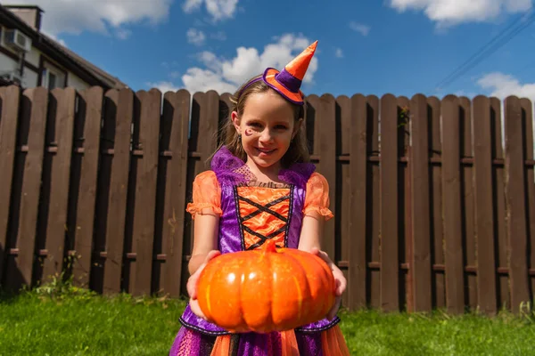 Criança em traje de palhaço e boné de festa segurando abóbora e sorrindo para a câmera — Fotografia de Stock