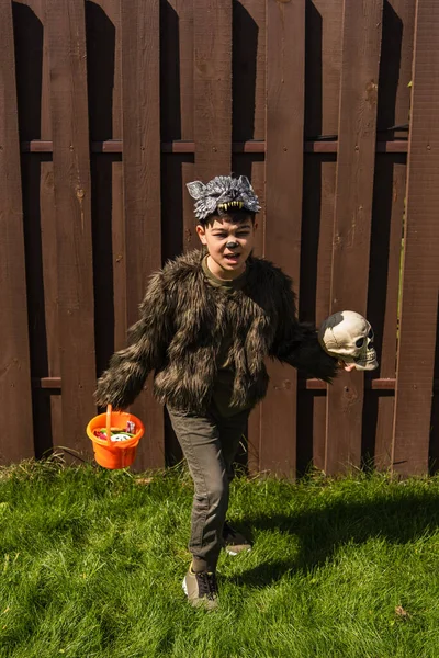 Pleine longueur de asiatique garçon en loup-garou costume courir avec crâne et halloween seau avec des bonbons dans arrière-cour — Photo de stock