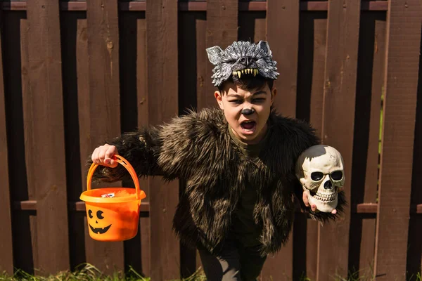 Asian boy in werewolf costume running with skull and trick or treat bucket while scary screaming outdoors — Stock Photo