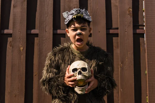 Asiático niño en hombre lobo traje gruñendo y sosteniendo cráneo mientras mirando cámara - foto de stock