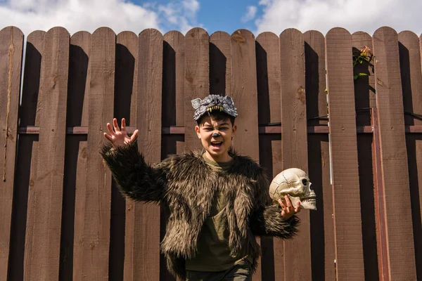 Asiatico ragazzo in lupo mannaro costume mostrando spaventoso smorfia e gesturing mentre holding spooky cranio — Foto stock