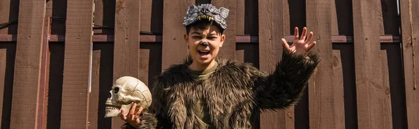 Garçon asiatique en costume de loup-garou montrant grimace en colère et geste effrayant tout en tenant le crâne effrayant, bannière — Photo de stock
