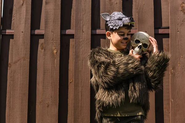 Feliz asiático chico en halloween hombre lobo traje abrazando espeluznante cráneo cerca de madera valla - foto de stock