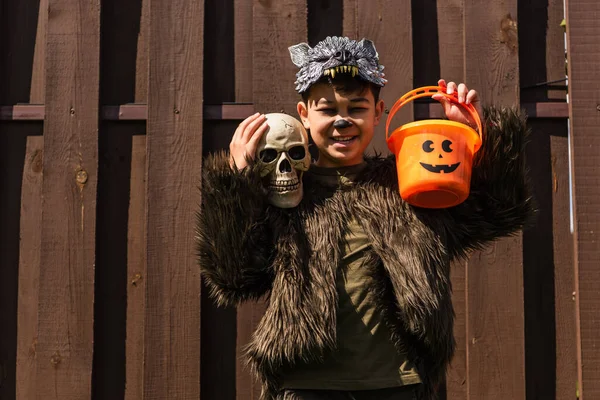 Allegro asiatico ragazzo in lupo mannaro costume holding cranio e halloween secchio — Foto stock