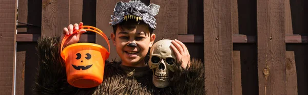 Asian boy in werewolf costume holding halloween bucket and skull while smiling at camera, banner — Stock Photo