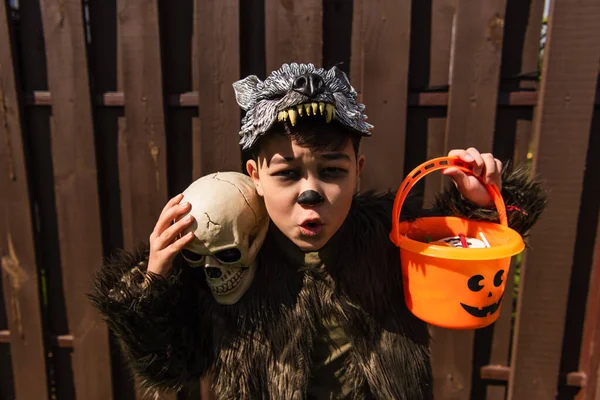 Excited asian boy in werewolf costume holding skull and halloween bucket with sweets — Stock Photo