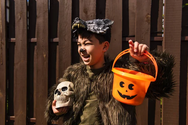 Excited asian boy in werewolf costume holding halloween bucket and spooky skull while looking away — Stock Photo