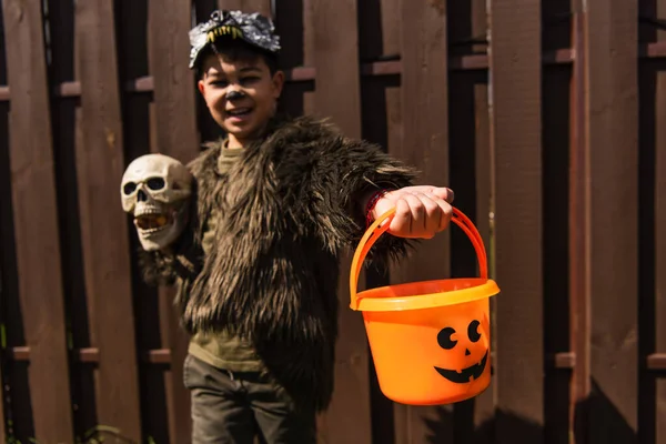 Asiático chico en hombre lobo traje celebración cráneo y halloween cubo mientras sonriendo en borrosa fondo - foto de stock