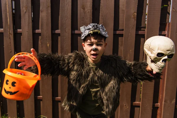 Asombrado asiático chico en hombre lobo traje corriendo con cráneo y halloween cubo en patio trasero - foto de stock