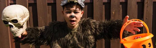 Asombrado asiático chico en hombre lobo traje celebración cráneo y halloween cubo con dulces, bandera - foto de stock