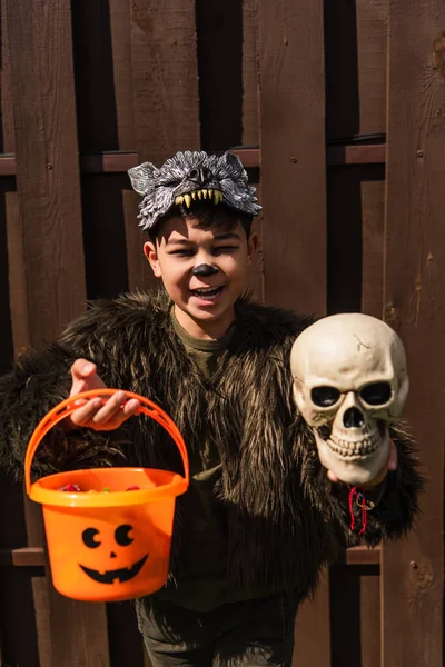 Happy asian boy in werewolf costume holding spooky skull and halloween bucket while looking at camera — Stock Photo