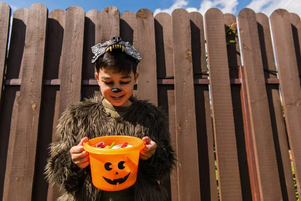 Pleased asian boy in werewolf costume looking into halloween bucket with sweets — Stock Photo