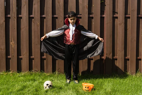 Full length of asian boy in vampire king costume holding cape near skull and halloween bucket on lawn — Stock Photo