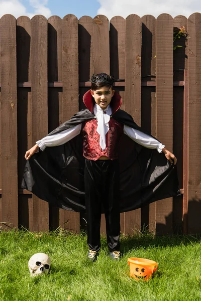 Pleine longueur de asiatique garçon dans vampire costume tenue manteau près crâne et halloween seau sur herbe — Photo de stock