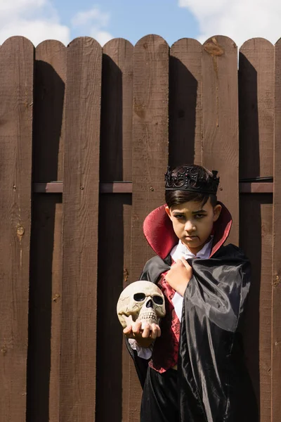 Serious asian boy in vampire costume wrapping in cape while standing with skull near wooden fence — Stock Photo