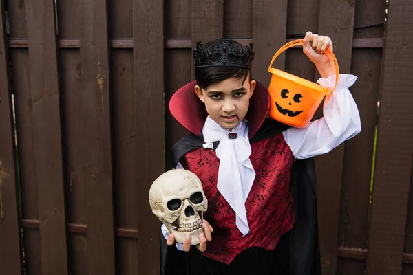 Asian boy in vampire costume holding skull and halloween bucket outdoors — Stock Photo