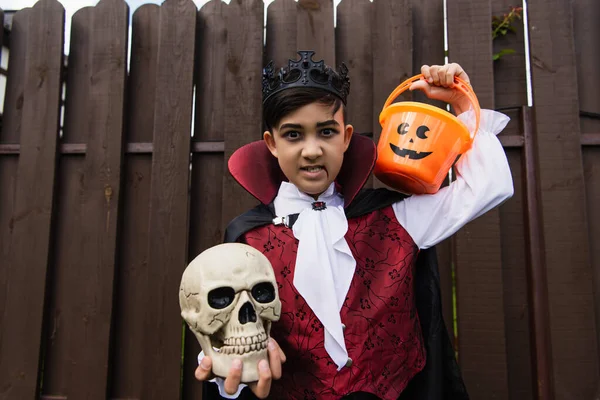 Asiático chico con enojado mueca usando vampiro traje y sosteniendo cráneo y truco o tratar cubo - foto de stock