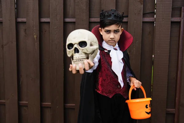 Serious asian boy in vampire costume holding halloween bucket and scary skull in outstretched hand — Stock Photo