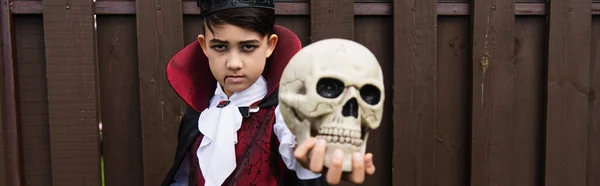 Serios asiático chico en vampiro traje celebración espeluznante cráneo y mirando cámara, bandera - foto de stock