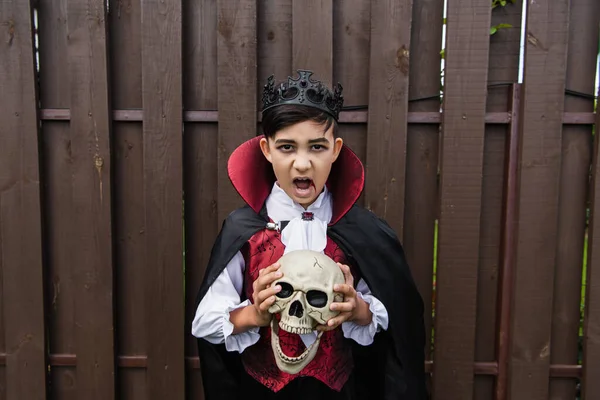 Asian boy in vampire costume holding skull and screaming while looking at camera — Stock Photo