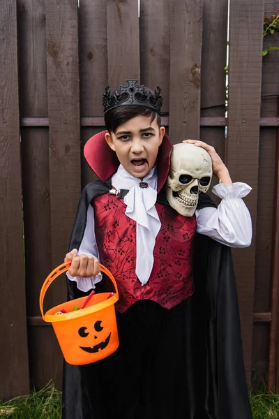 Asiático chico en halloween traje mostrando enojado mueca y gritando con cráneo y truco o tratar cubo - foto de stock