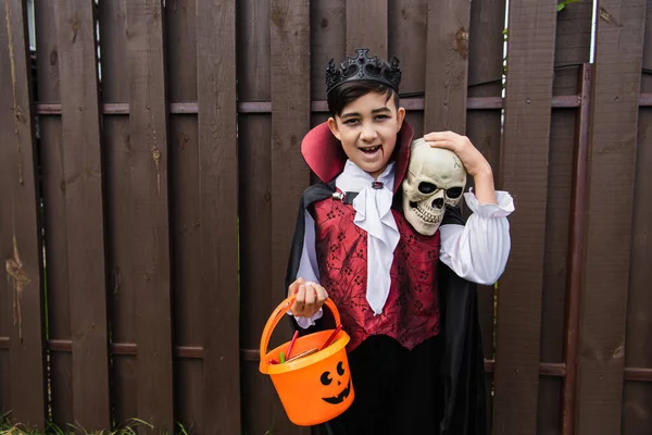 Feliz asiático chico en vampiro traje celebración cráneo y truco o tratar cubo mientras mira cámara - foto de stock