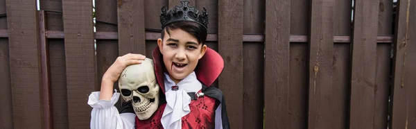 Glücklicher asiatischer Junge im Vampirkostüm schaut in die Kamera, während er Totenkopf, Banner hält — Stockfoto