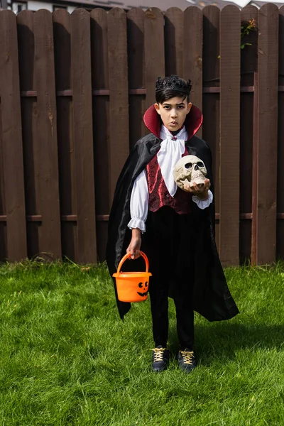 Asiático chico en vampiro rey traje y enojado mueca celebración halloween cubo y cráneo - foto de stock