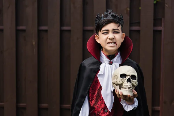 Ásia menino no vampiro rei traje rosnando enquanto segurando crânio e olhando para câmera — Fotografia de Stock