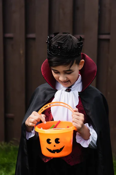 Asiático niño en vampiro rey traje buscando en truco o tratar cubo con caramelos - foto de stock