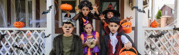 Девушки, показывающие страшные жесты рядом с многонациональными друзьями в костюмах на Хэллоуин, баннер — стоковое фото